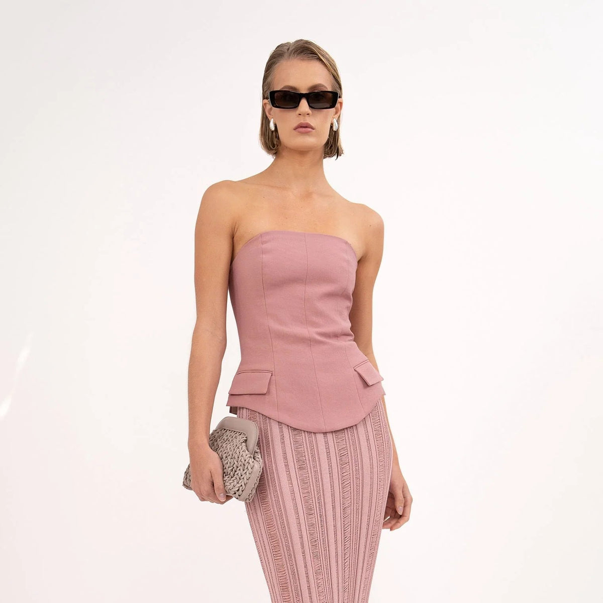 Woman wearing a pink strapless top and pleated pants with sunglasses on a white background