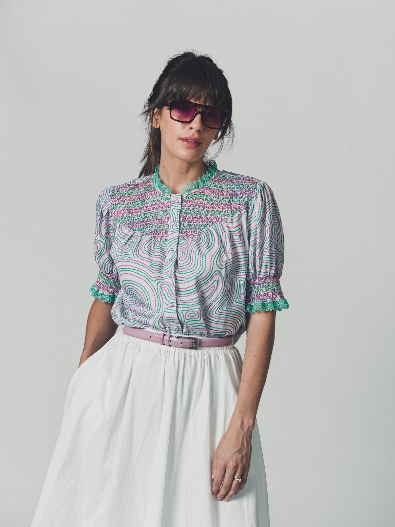 Model wearing a cropped cotton blouse with puff sleeves, smocked detailing, and green maze print, styled with a white gathered skirt and oversized sunglasses.