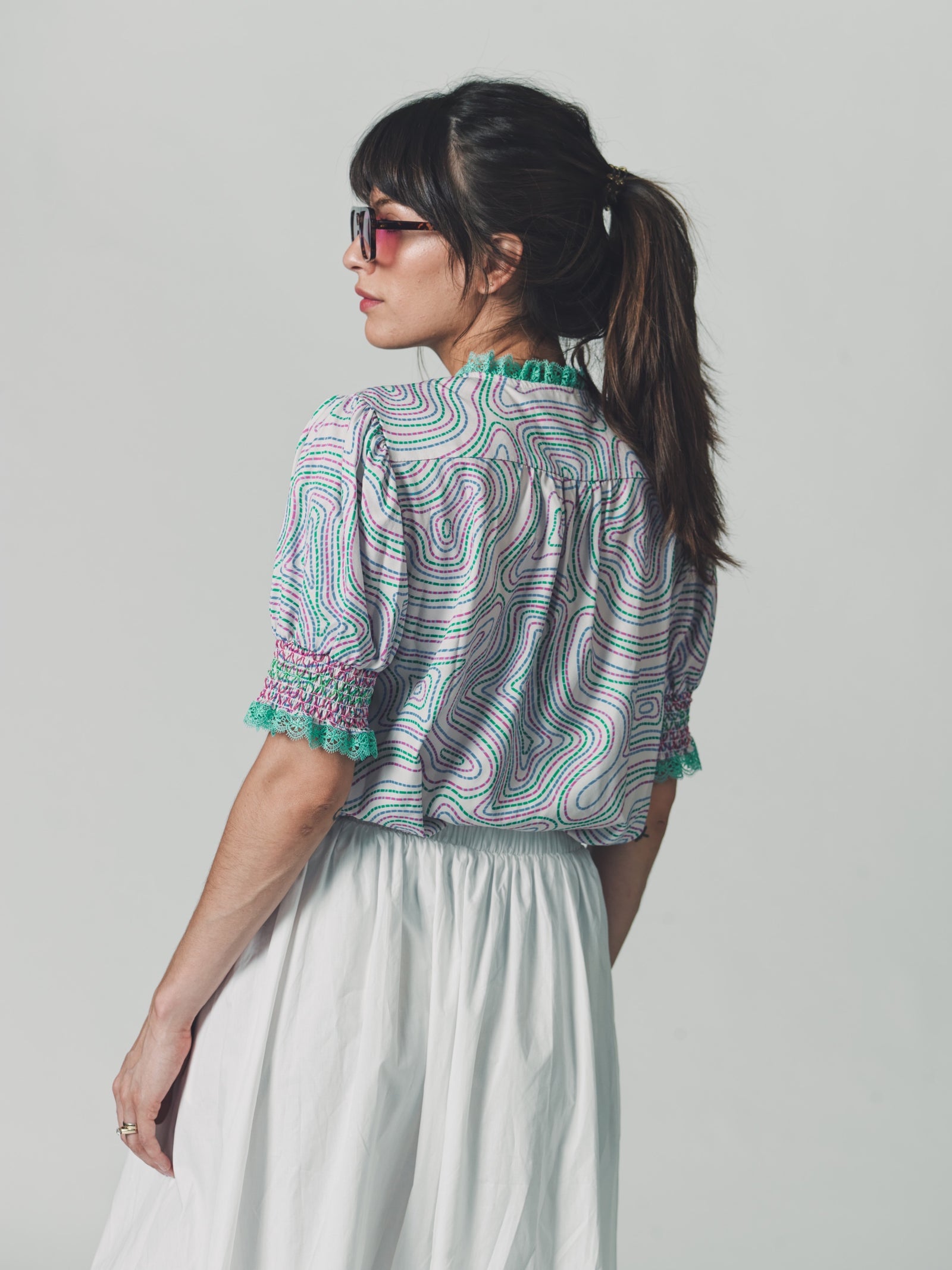 Model wearing a cropped cotton blouse with puff sleeves, smocked detailing, and green maze print, styled with a white gathered skirt and oversized sunglasses.