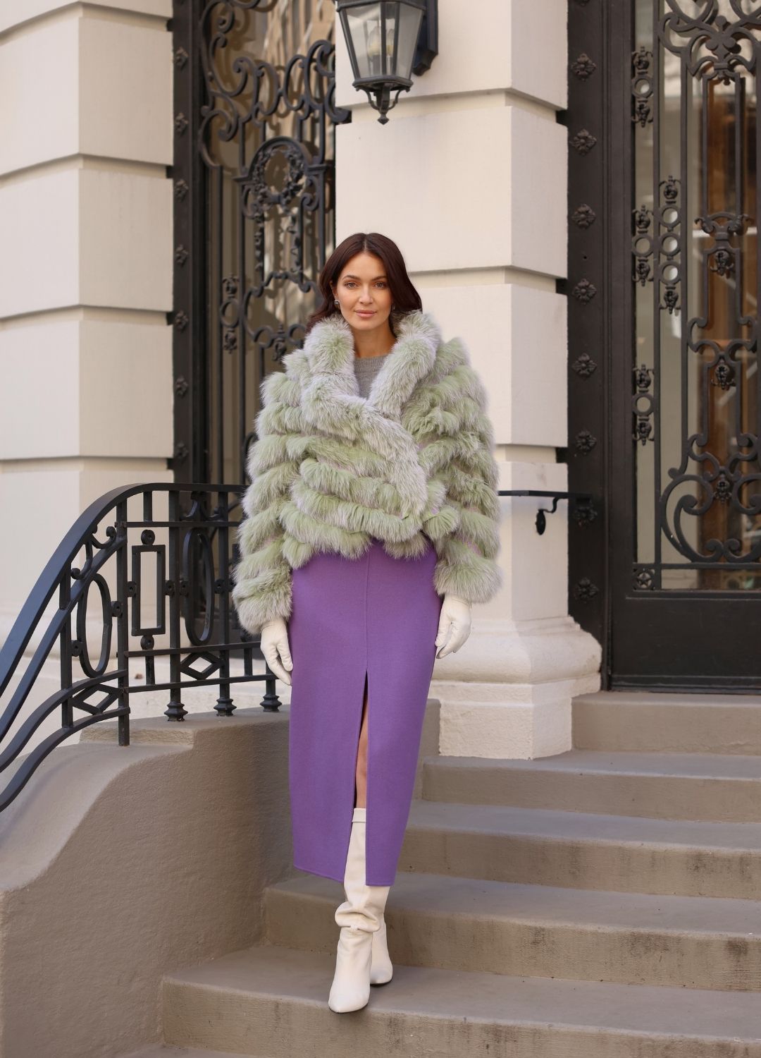 Woman in a green fur coat and purple skirt standing on steps in an urban setting
