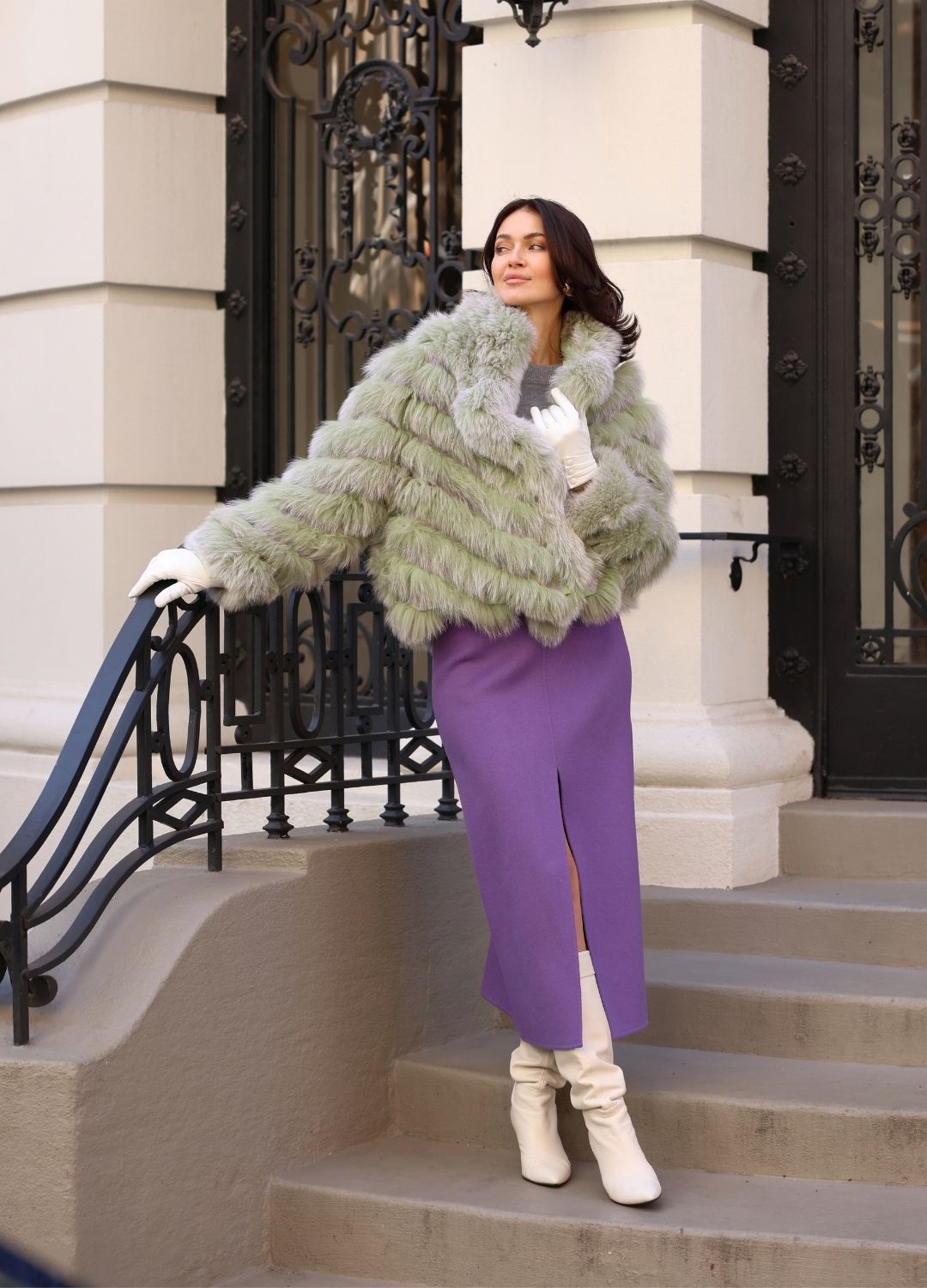 Woman in a green fur coat and purple pants standing on steps outside a building.