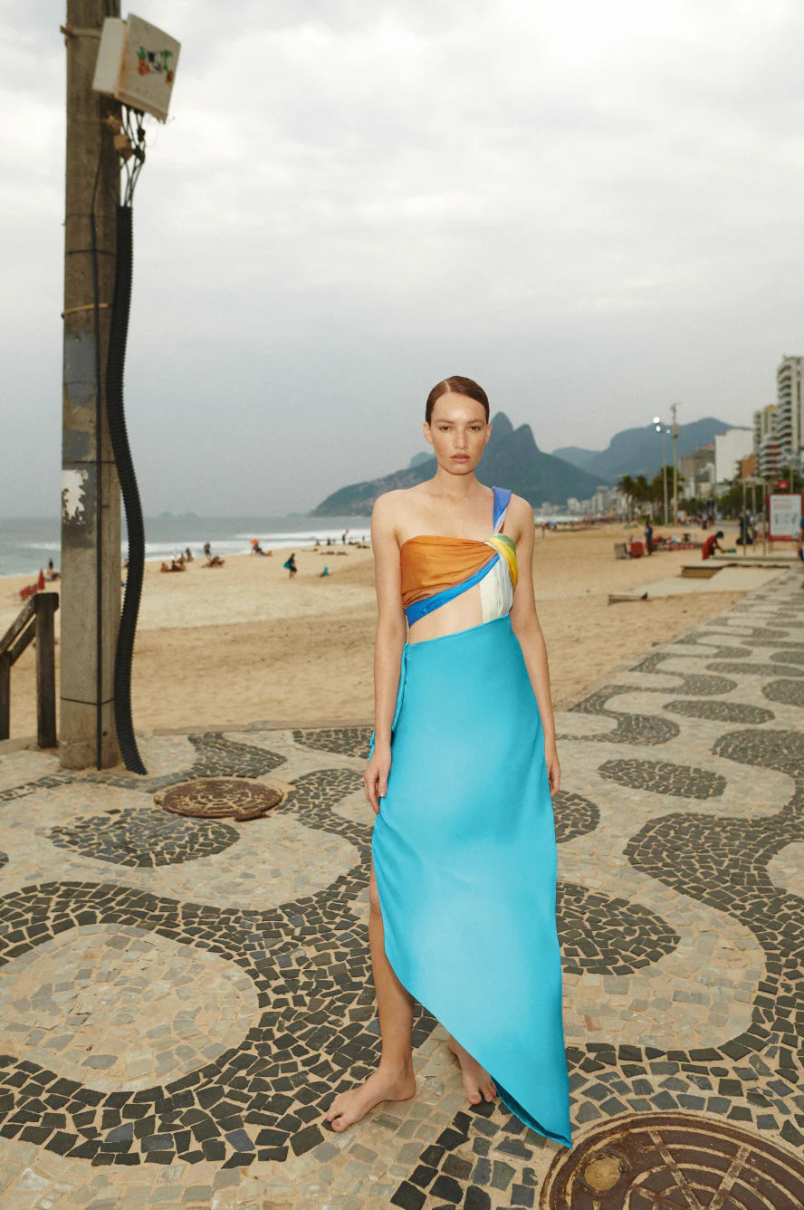 Woman in a blue and brown dress standing on a beach with mountains in the background