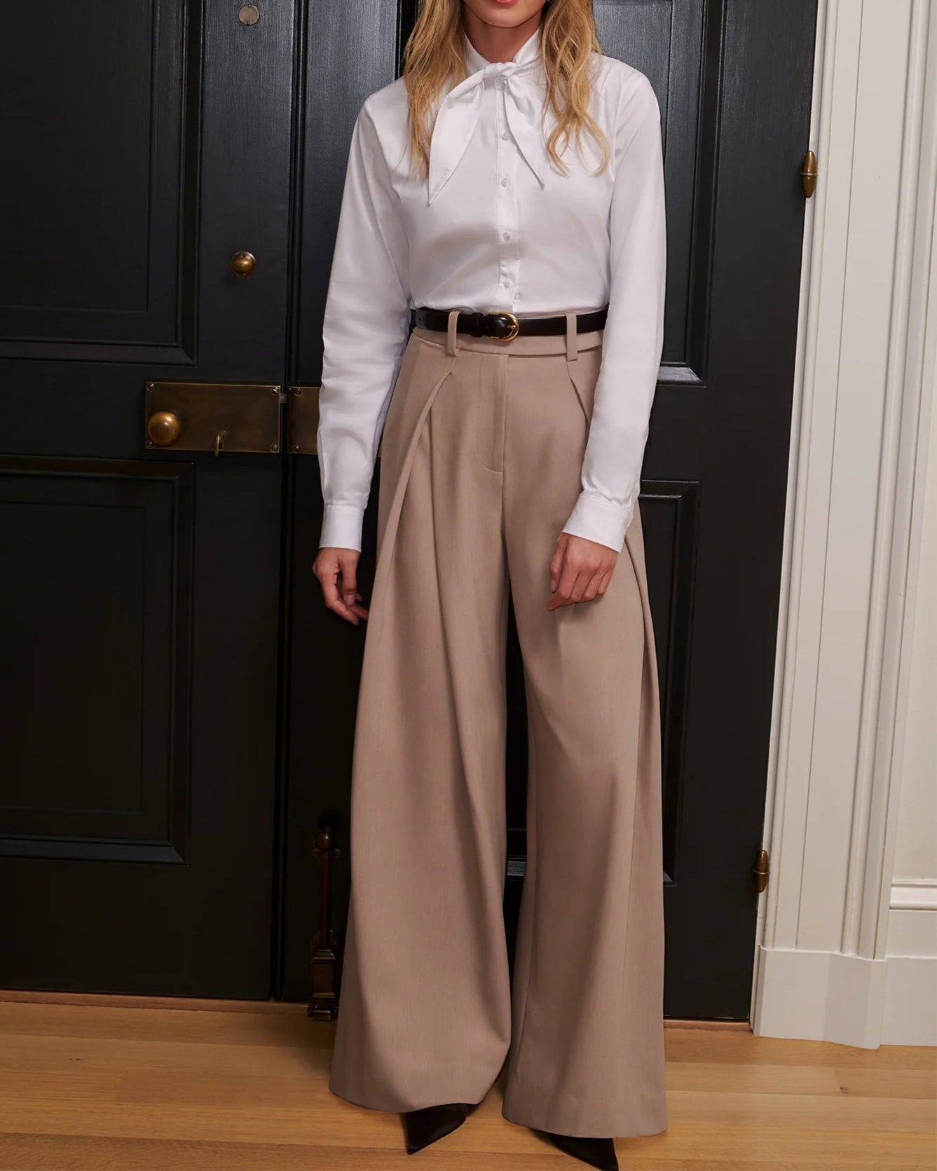 Woman wearing a white blouse and beige wide-leg pants standing in front of a dark door.